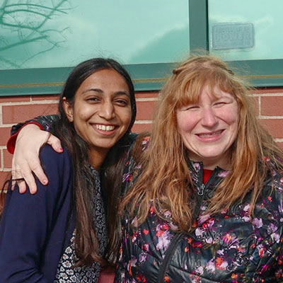 Two smiling, young women leaning into each other, with one woman’s arm on the other’s shoulder.