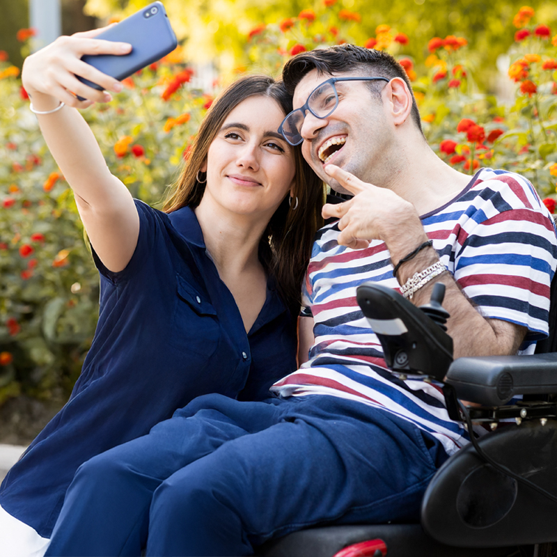 A man between the ages of 30 and 40 with a disability sits in an automatic wheelchair outdoors with a female caregiver.The couple takes a selfie with their mobile phone.